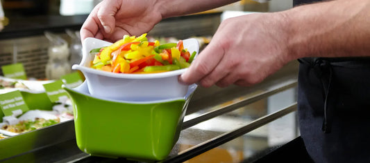 Close-up of a server placing a white bowl of freshly sliced red, yellow, and green peppers into a larger green display container. Other colorful prepared dishes in matching containers are visible blurred in the background.