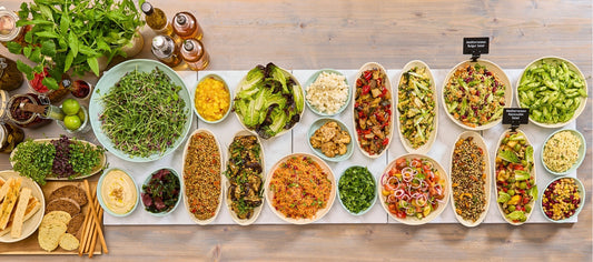 Overhead salad bar display featuring a wide variety of fresh dishes including grains, vegetables and Mediterranean-style salads arranged in ceramic serving bowls on a clean, neutral counter.