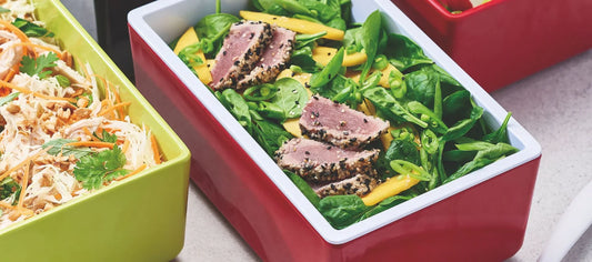 Close-up of a rectangular red tray filled with a fresh salad of seared sesame-crusted tuna slices, spinach leaves, mango strips, and green beans. A lime-green tray with shredded chicken and carrots sits beside it.