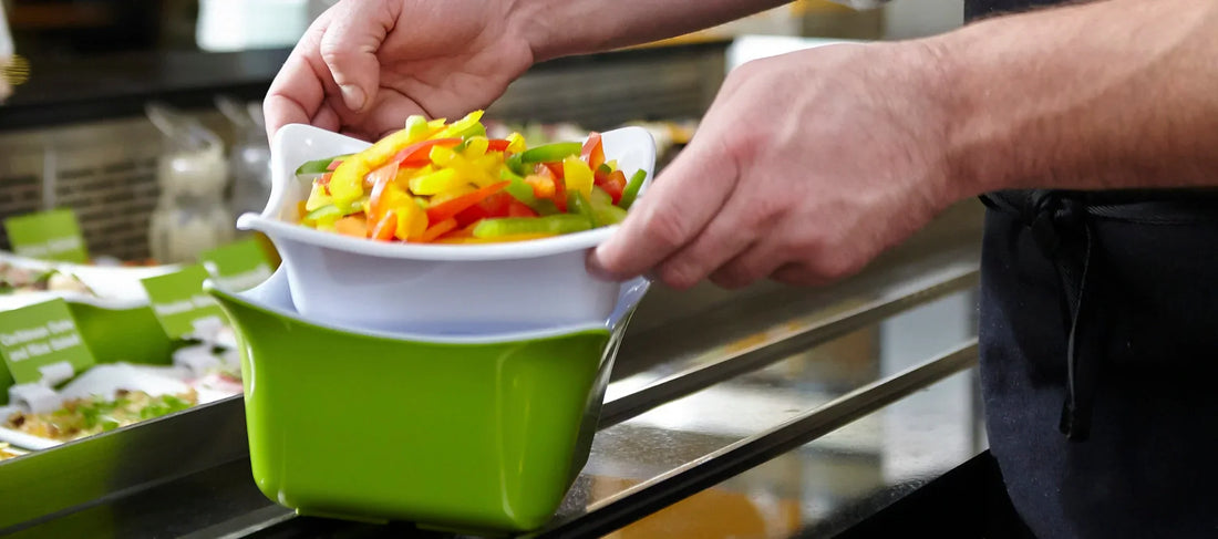 Close-up of a server placing a white bowl of freshly sliced red, yellow, and green peppers into a larger green display container. Other colorful prepared dishes in matching containers are visible blurred in the background.