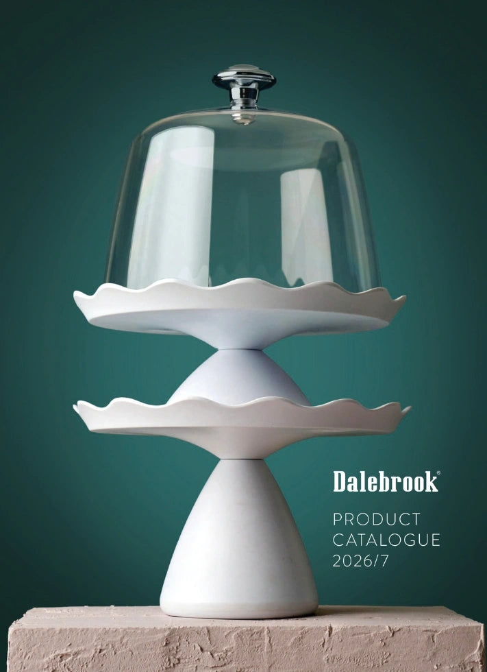 A white two-tier pedestal cake stand with scalloped edges under a clear glass dome, shown on a textured stone surface against a dark teal background. White text on the right reads 'Dalebrook Product Catalogue 2026/7'.