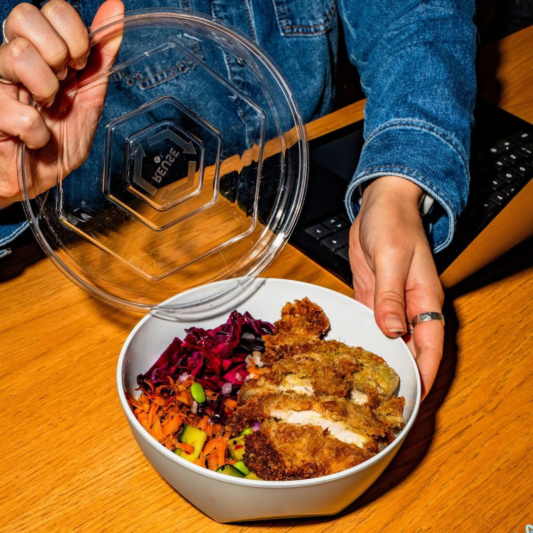 Person holding a clear reusable lid over a reusable bowl of food on a wooden desk beside a laptop