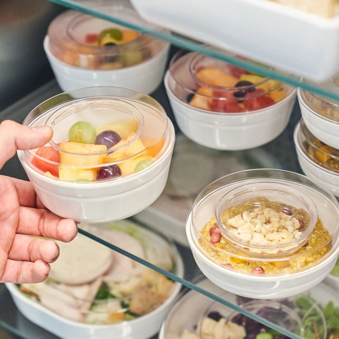 Hand holding a small container of fruit salad with a display case in the background showing various pre-made meals in packaged in reusable bowls