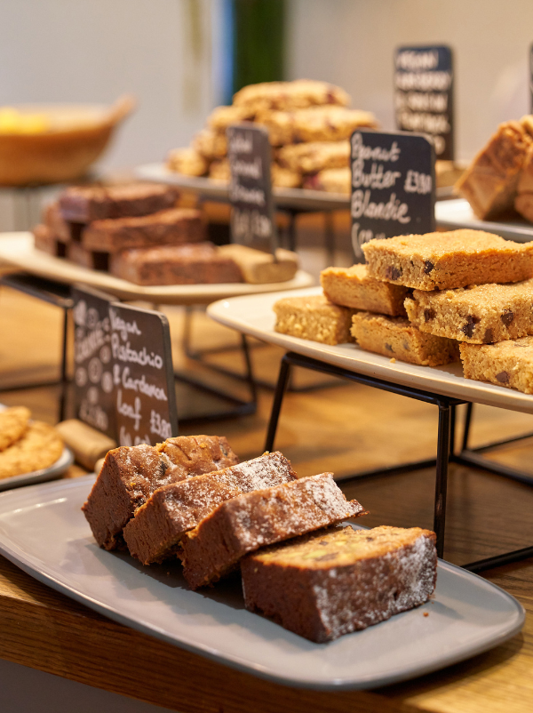 A bakery display with assorted baked goods on white trays and black metal stands, including brownie bars, scones, and cake slices, with handwritten price signs on a wooden counter.