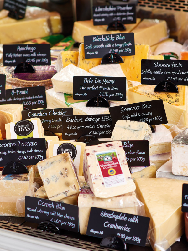 A display of assorted cheese wedges and blocks in a market setting, each with a handwritten black price tag card. Visible labels include Blackstick Blue, Brie De Marnier, Canadian Cheddar, Somerset Brie, Cornish Blue, and Wendylecde with Cranberries.