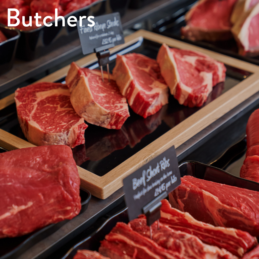 Raw beef cuts displayed on black trays in a butcher shop counter, with wooden trays holding marbled red steaks and red meat slices in the foreground. Small price tags are visible, and the word 'Butchers' appears in white text at the top left.
