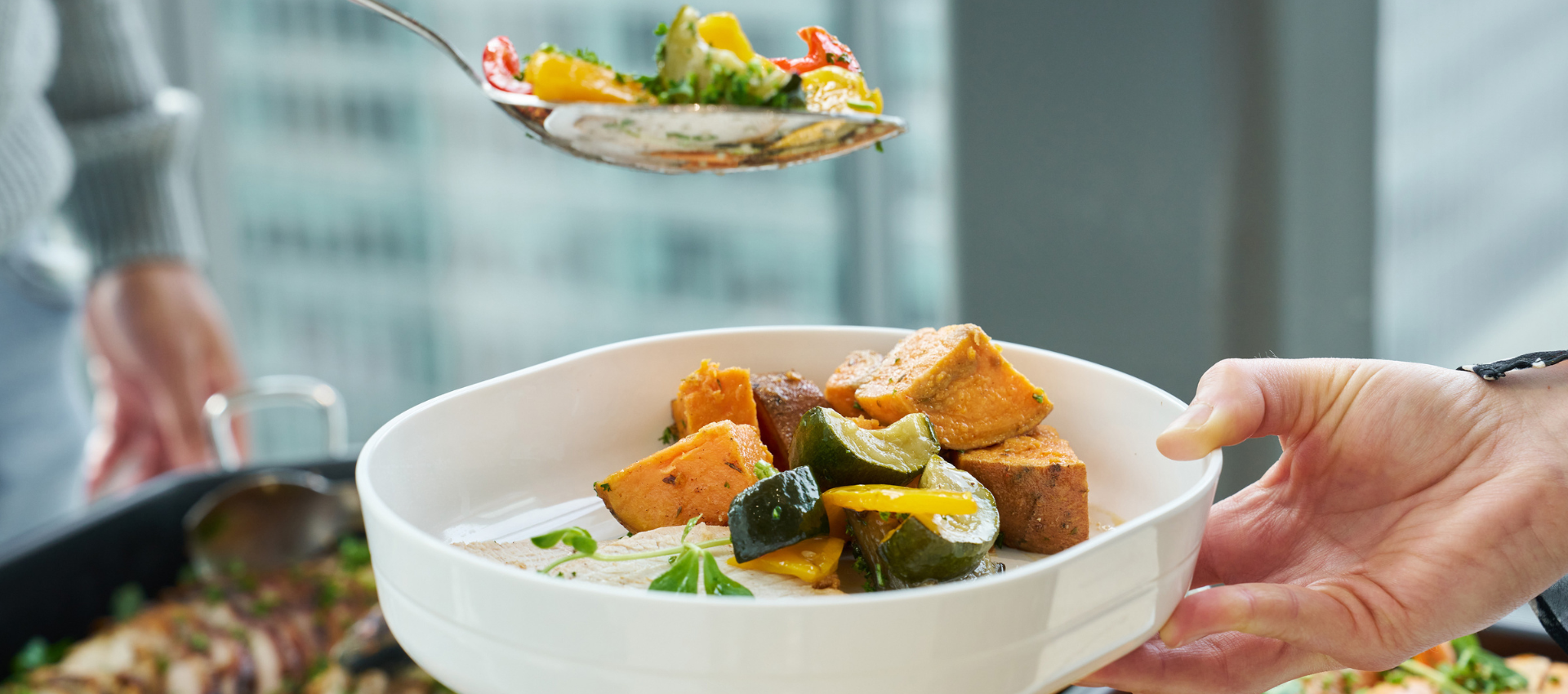 A close-up of a white ceramic bowl being held by a hand, filled with a colorful vegetable and sweet potato dish, with another hand holding a spoon above it. The background shows a softly blurred kitchen or dining environment with a window and city view.
