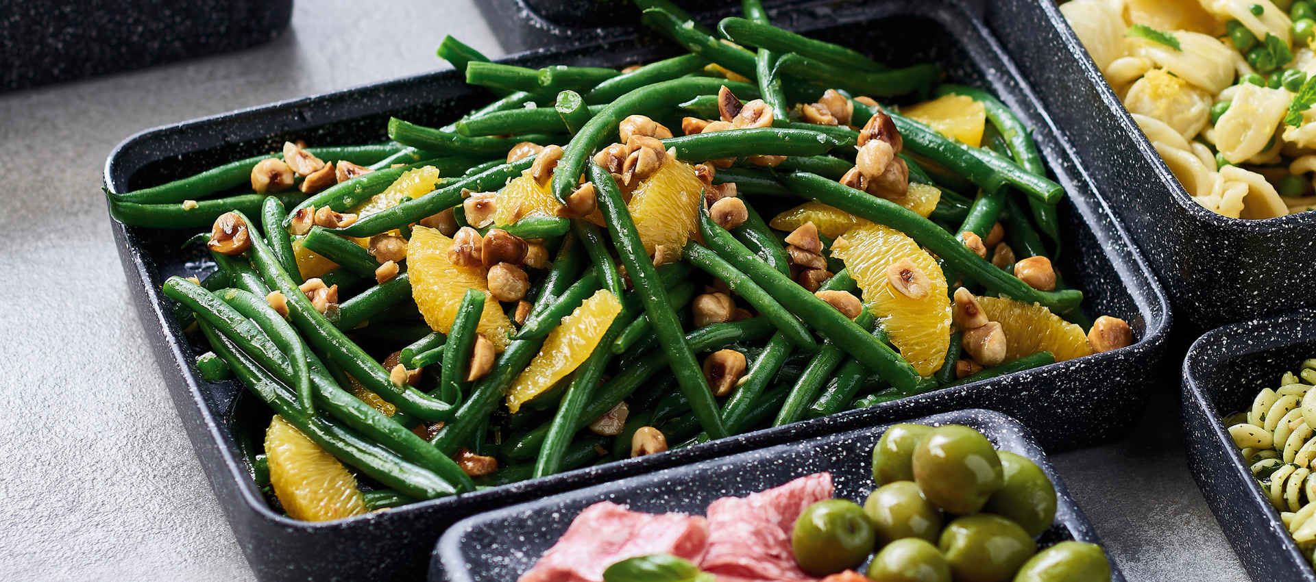 A close-up of a black speckled rectangular tray filled with cooked green beans, orange slices, and chopped nuts, set on a light gray surface with other food trays partially visible around it.