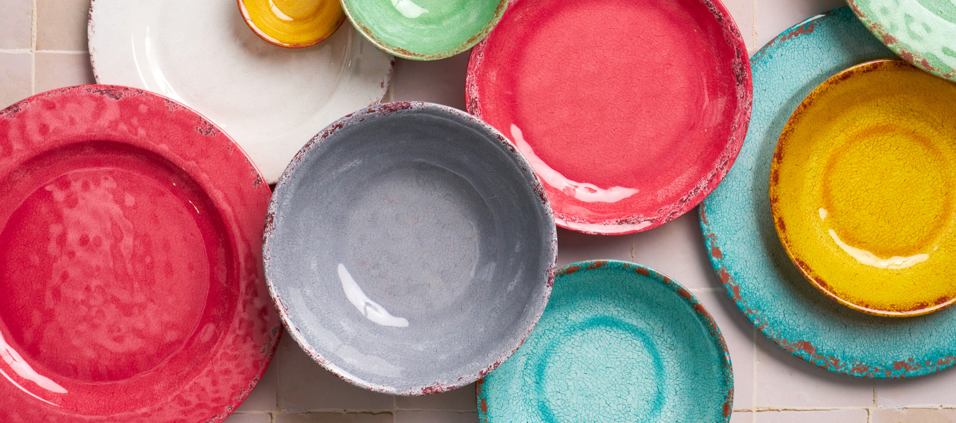 A collection of colorful handmade ceramic plates and bowls with a distressed, rustic finish, arranged on a light tiled surface.