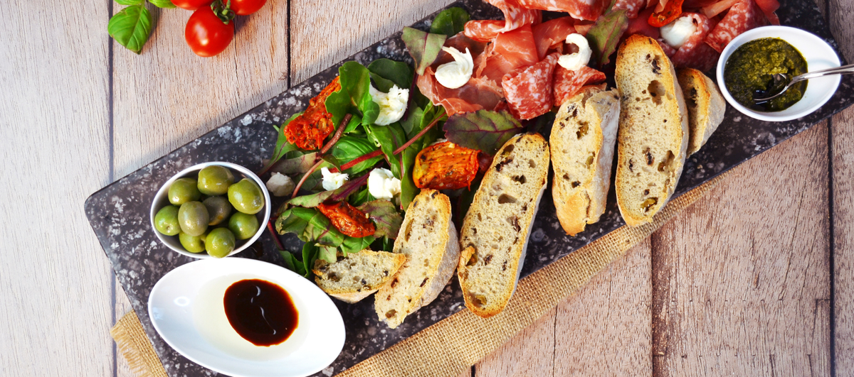 An overhead styled food platter on a dark stone serving board, featuring sliced toasted bread, greens, prosciutto, white cheese, olives, a small bowl of green sauce, and a small bowl of dark balsamic. The platter is set on a rustic light wood surface.