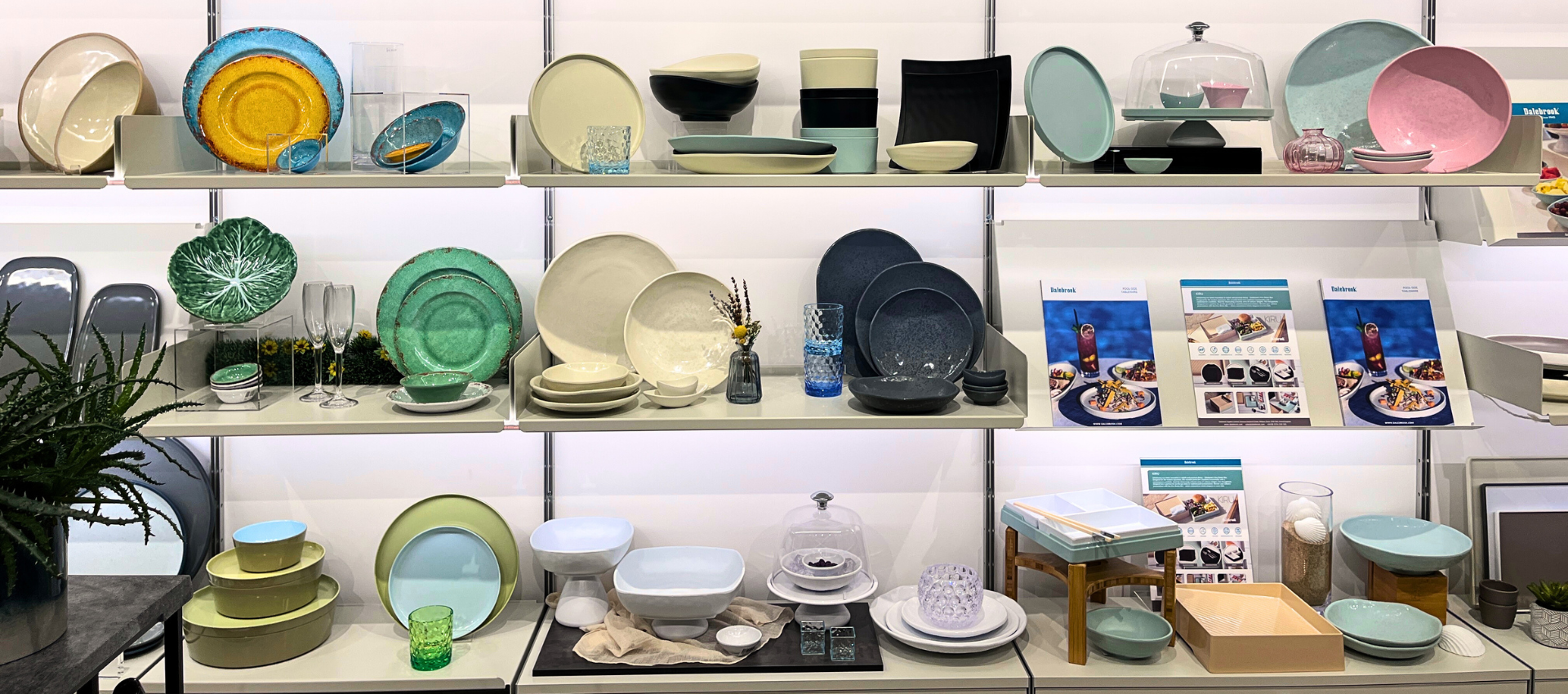 A display of Dalebrook tableware and serving dishes arranged on multiple white shelves in a showroom. The collection includes plates, bowls, casserole dishes, and small serving pieces in pastel and earthy tones such as mint green, pale blue, pink, cream, and dark charcoal.