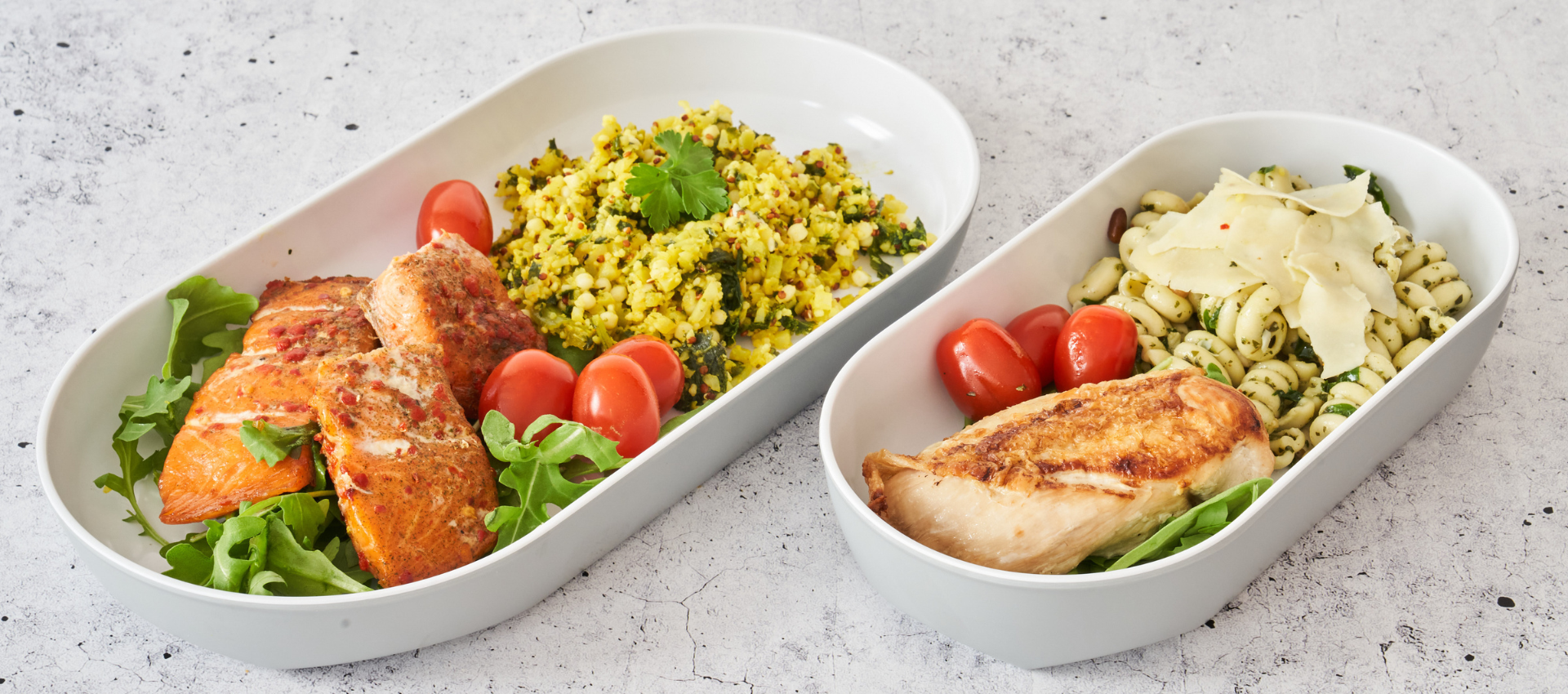 Two white oval meal prep containers filled with prepared food on a light stone surface. The left container holds grilled salmon, yellow rice, cherry tomatoes, and greens.