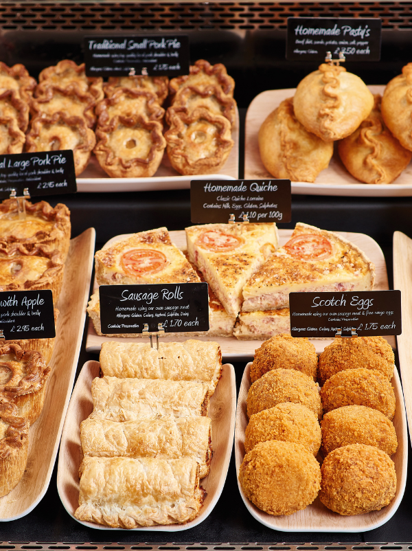 A bakery display case filled with assorted baked goods and savory pastries, including mini pies, sausage rolls, scotch eggs, and homemade quiches, each with handwritten price tags on black labels.