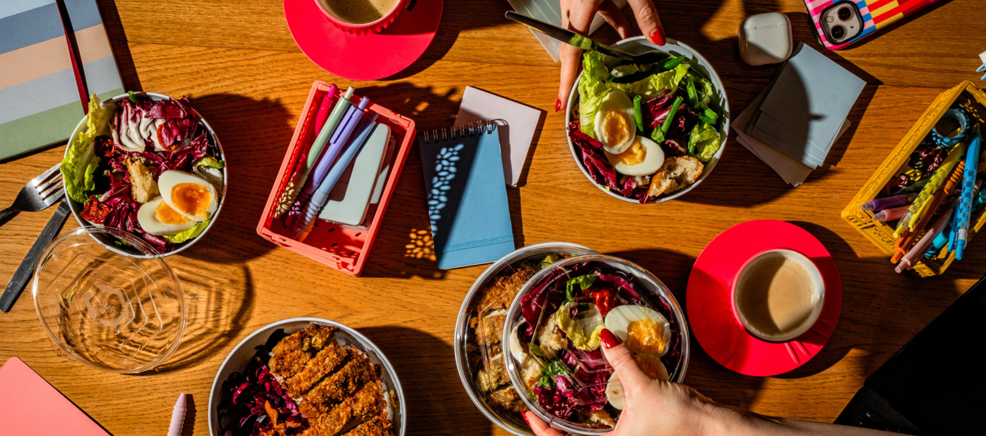 Overhead view of a wooden table styled with bowls of salad, breakfast items, colorful notebooks, and red saucers, with hands reaching in to grab food.