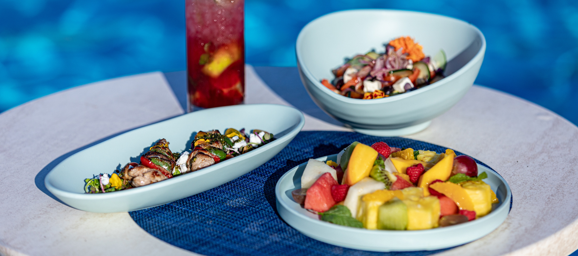 Three light blue ceramic dishes containing a colorful salad, a fruit skewer, and a prepared dish, arranged on a round table with a blue textured placemat and a glass of red drink in the background.