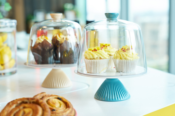 A set of clear glass cupcake domes and cake stands on a white table, with cupcakes under a blue stand in sharp focus and other stands blurred in the background.
