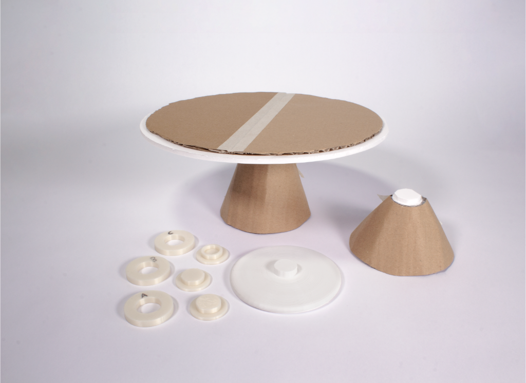 A handmade cardboard and white plastic cake stand or pedestal set, shown with its separate base, top disc, small cone-shaped piece, and several round white plastic inserts on a light gray studio background.