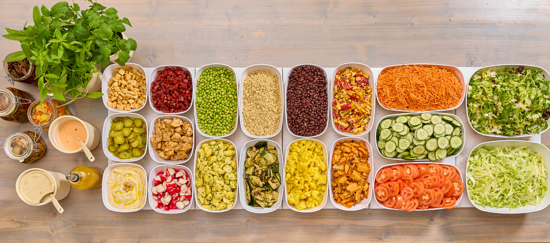 A top-down view of a large salad bar or buffet spread on a wooden table, featuring multiple white trays filled with various colorful vegetables, grains, beans, and dressings.