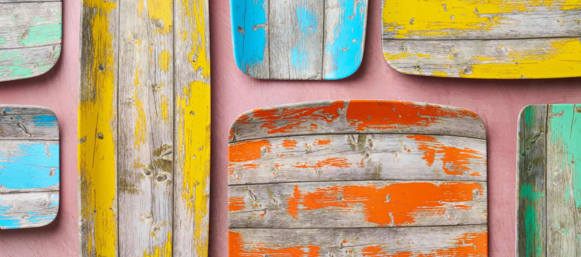 collection of rustic wooden serving boards with distressed painted finishes in yellow, orange, turquoise, and green, mounted on a soft pink wall.