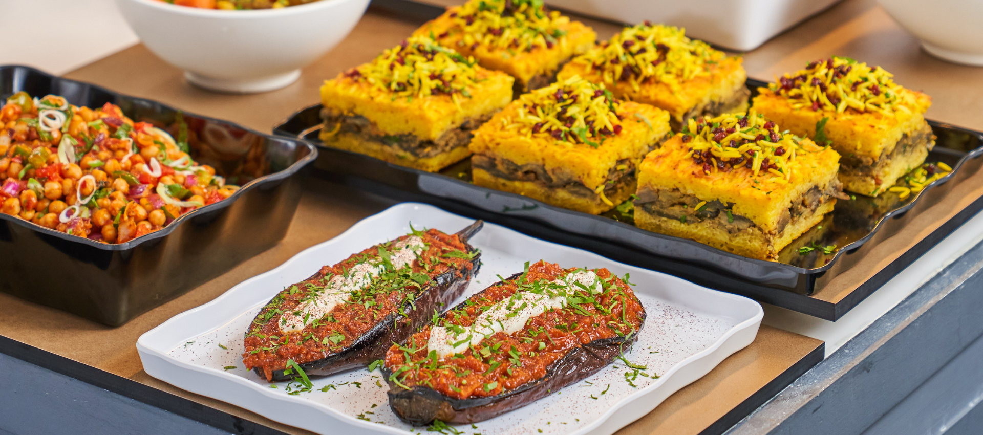 A close-up of prepared food on a buffet or serving table, featuring stuffed eggplant slices topped with red sauce and herbs on a white plate, and several yellow-topped layered dessert or casserole squares on a black tray.